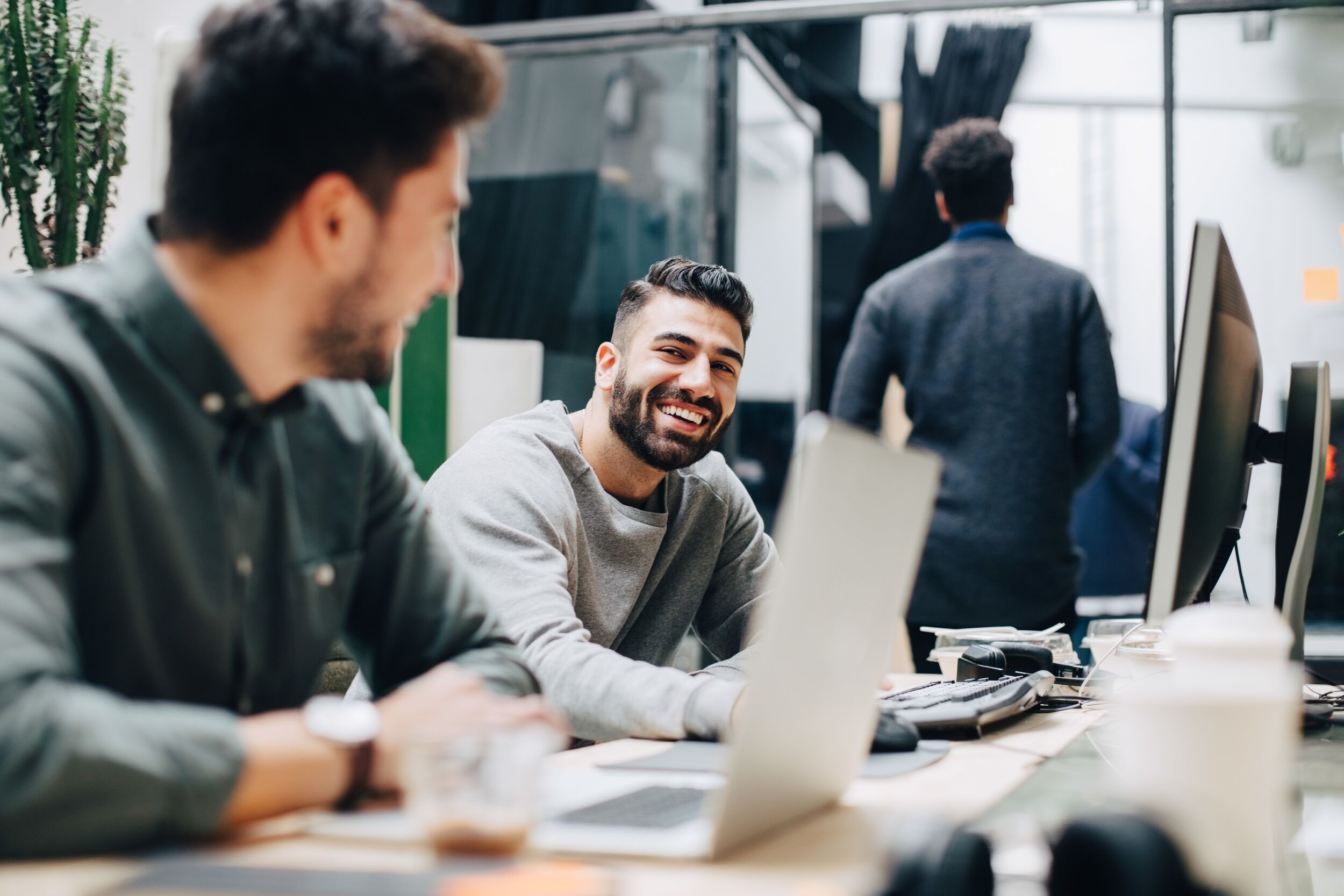 Smiling male colleagues looking at each other while sitting by desk in office.