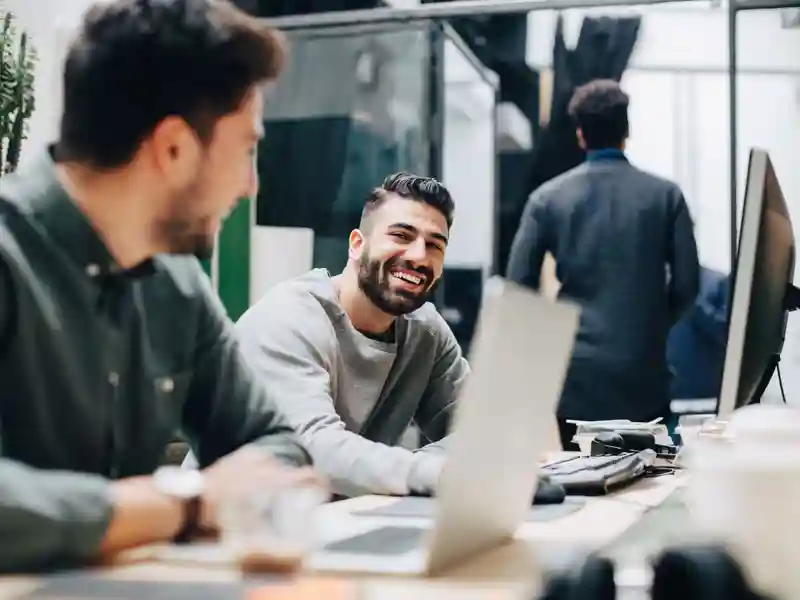 Smiling male colleagues looking at each other while sitting by desk in office.